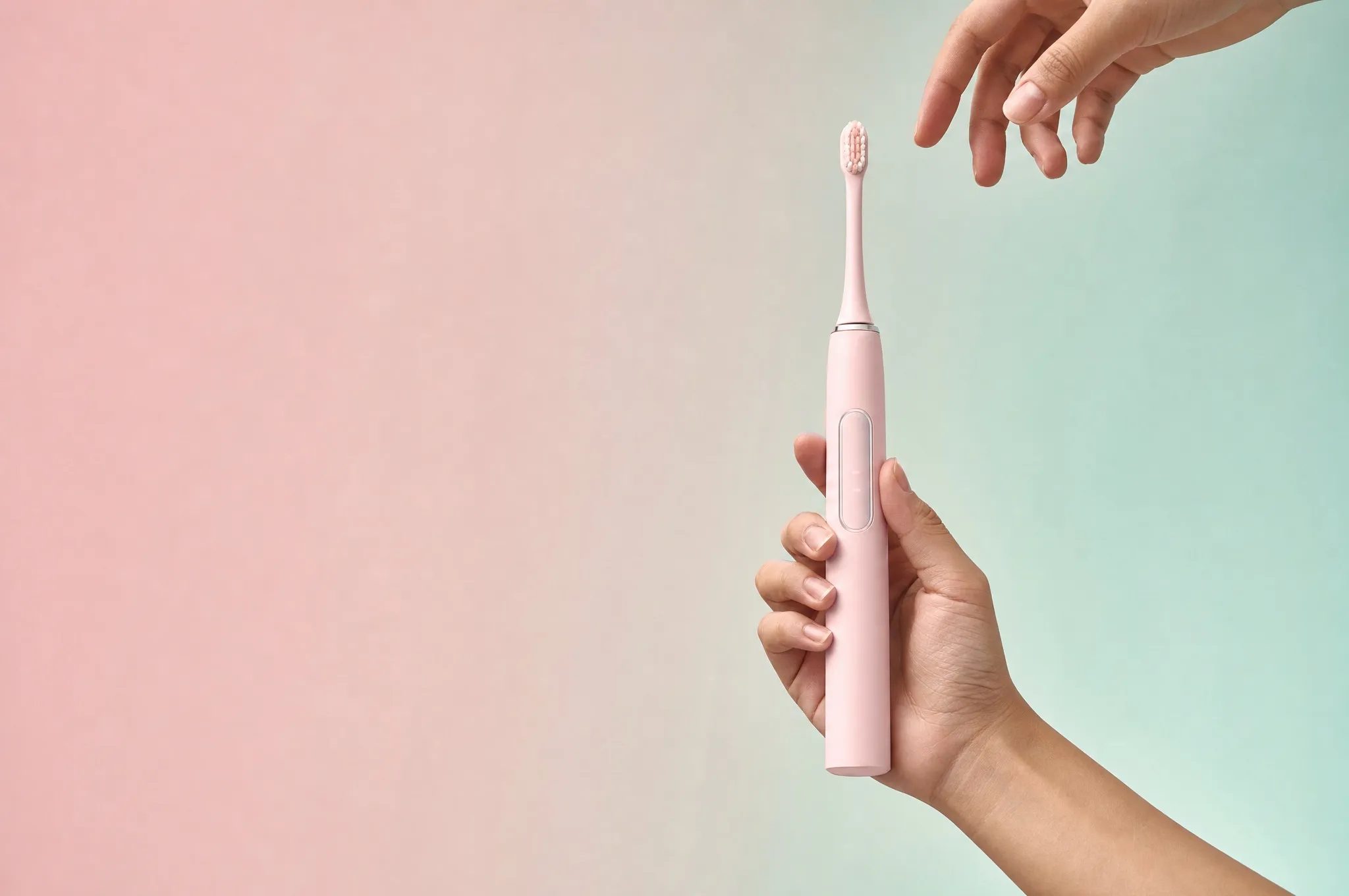 A hand holding a pink electric toothbrush, a second hand reaching toward it