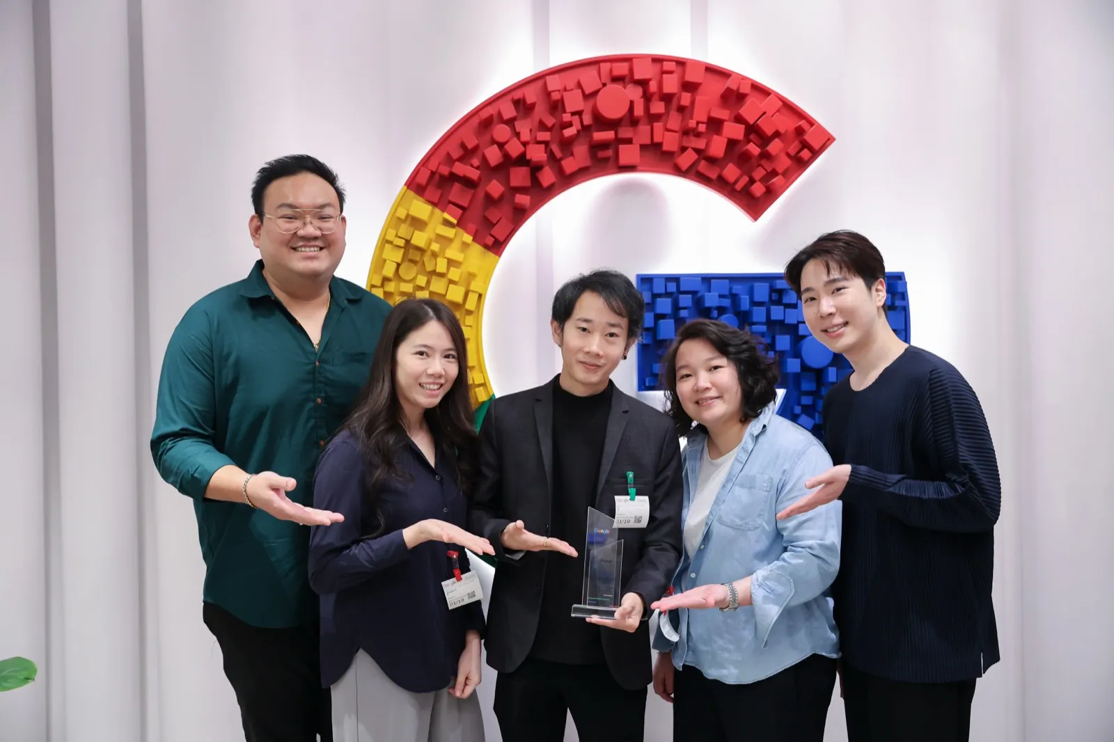 Five Primal team members wearing event lanyards, smiling around a trophy in front of a large multicoloured letter G wall