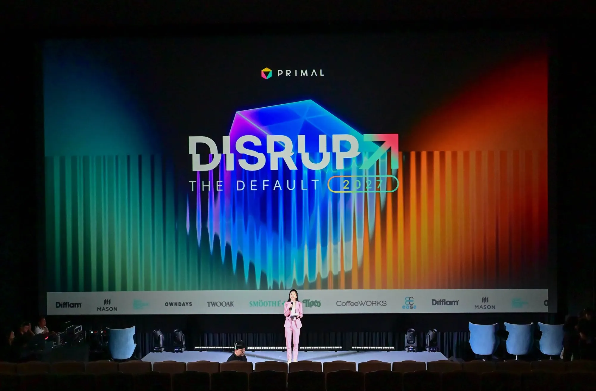 A speaker on stage in front of the Disrupt The Default 2027 screen at Primal’s event