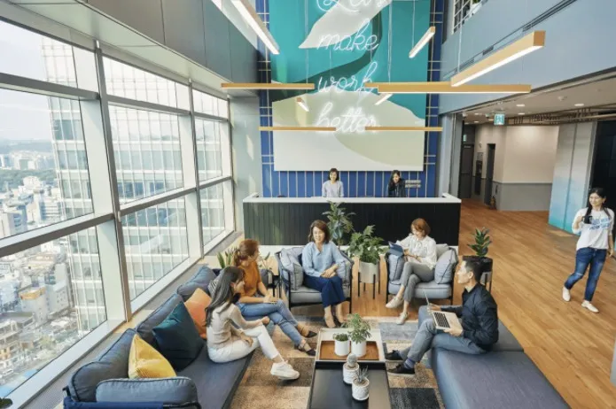 A JustCo coworking lounge: people talking on sofas beside floor-to-ceiling windows over a city, a reception desk under a neon sign behind them